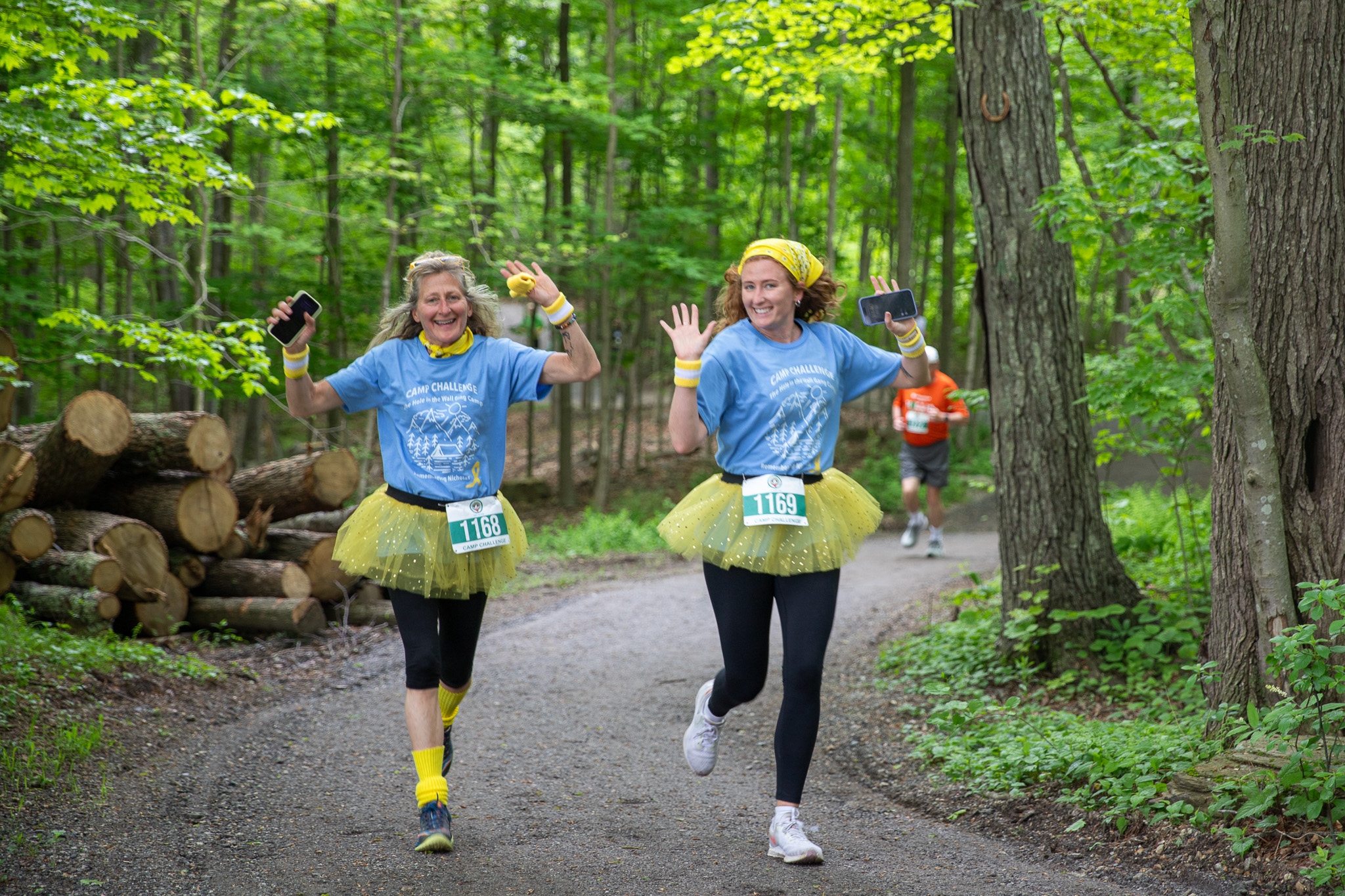 Two women in yellow tutus smile and wave while running at Camp