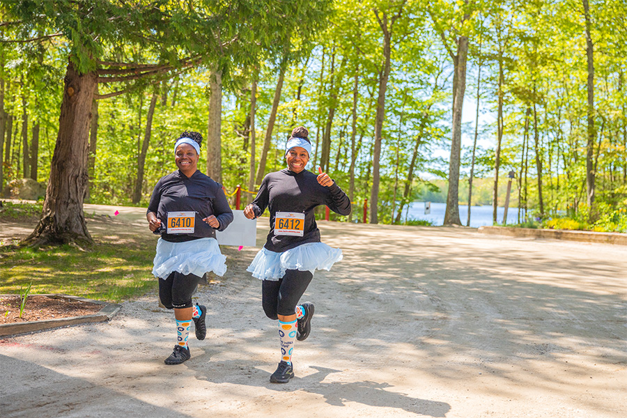 Two runners in blue tutus smile while running on the trails of Camp at the Connecticut Location