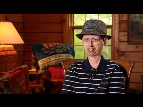 A person wearing glasses, a striped polo shirt, and a gray hat sits in a cozy wooden room, with a lamp and a colorful blanket in the background.