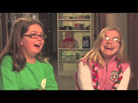 Two girls with glasses, smiling and sitting indoors. One wears a green shirt, the other wears a white top with a red vest. Shelves with books and toys are visible in the background.