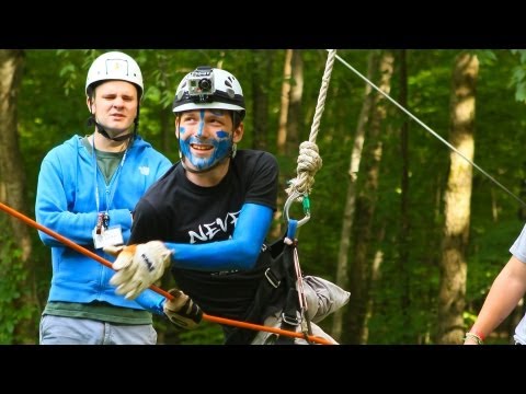 Two people wearing helmets and harnesses stand in a wooded area. The person in front, with blue face paint and gloves, is smiling and holding onto a rope, appearing ready to zipline.
