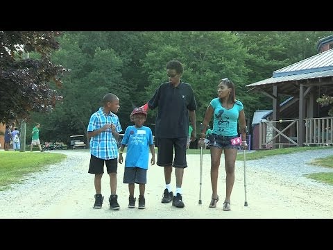 Four children walk on a gravel path outdoors; one uses crutches. Trees and a building with a porch are in the background. The children appear to be talking and smiling together.