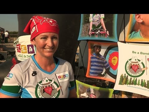 A woman in a red bandana and camp T-shirt smiles outdoors beside a display board featuring photos of camp activities and a sign that reads Be a Band..