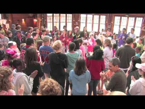 A large group of people, including children and adults, gather in a circle indoors, clapping and interacting in a lively, communal atmosphere. Sunlight streams through the windows in the background.