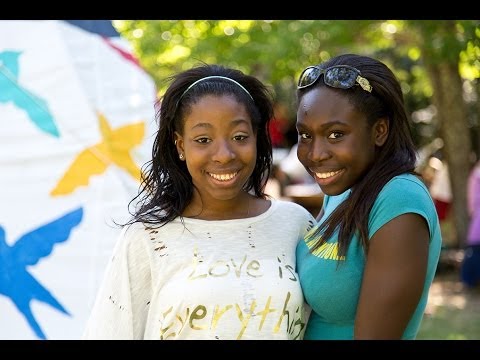 Two teenage girls smile together outdoors. One wears a white shirt with text and a headband, the other wears a teal shirt and sunglasses on her head. Colorful bird designs are visible in the background.