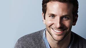 A man with short brown hair and blue eyes smiles at the camera, wearing a gray sweater over a blue-collared shirt, with a light blue background.