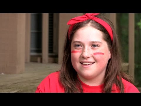 A smiling girl with long brown hair, wearing a red headband and red shirt, has two red stripes painted on each cheek. She is sitting outside near a wooden structure.