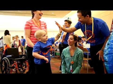 A smiling child with a bald head high-fives a man in a blue shirt. They are surrounded by happy adults, a wheelchair, and other people in a bright, indoor setting.