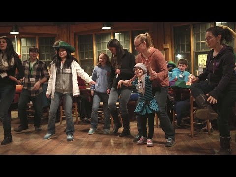 A group of people, including children and adults, are dancing together indoors on a wooden floor, with others sitting and watching in the background.