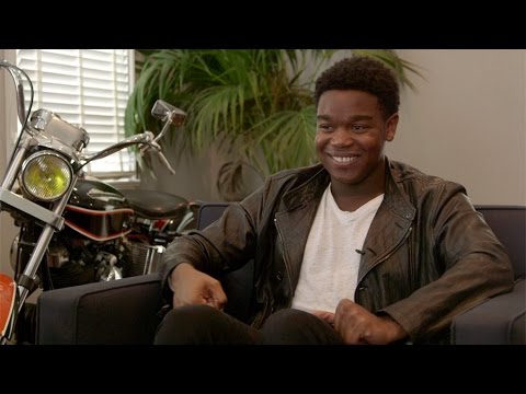 A smiling person in a black leather jacket sits on a dark sofa. There is a motorcycle and a green plant in the background. Light comes in through blinds on the left.