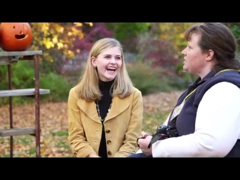Two women sit outdoors on a fall day, smiling and talking. One holds a camera, and a carved pumpkin sits on a wooden structure nearby. Colorful autumn leaves are visible in the background.