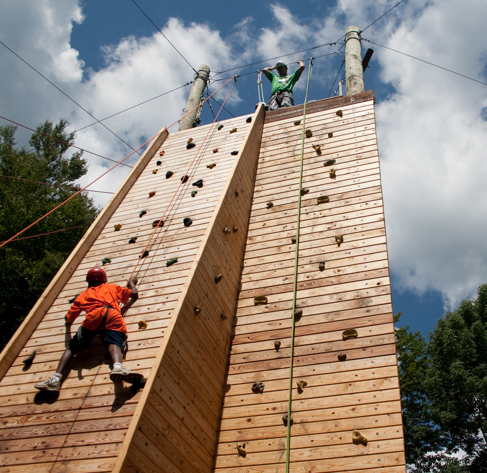 Two people climbing a tall outdoor wooden rock climbing wall on a sunny day, with one person near the top and another partway up. Trees and a blue sky with clouds are in the background.