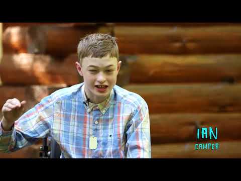 A teenage boy in a plaid shirt sits in front of a log cabin wall, smiling and gesturing with his hand. Text in the corner reads IAN CAMPER..