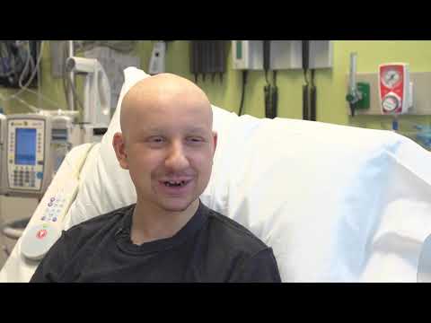 A person with a bald head sits up in a hospital bed, smiling. Medical equipment and monitors are visible in the background, suggesting a healthcare setting.