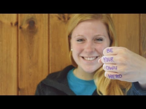Smiling person holds out a fist with BE YOUR OWN HERO written across their fingers in purple ink, standing in front of a wooden wall.