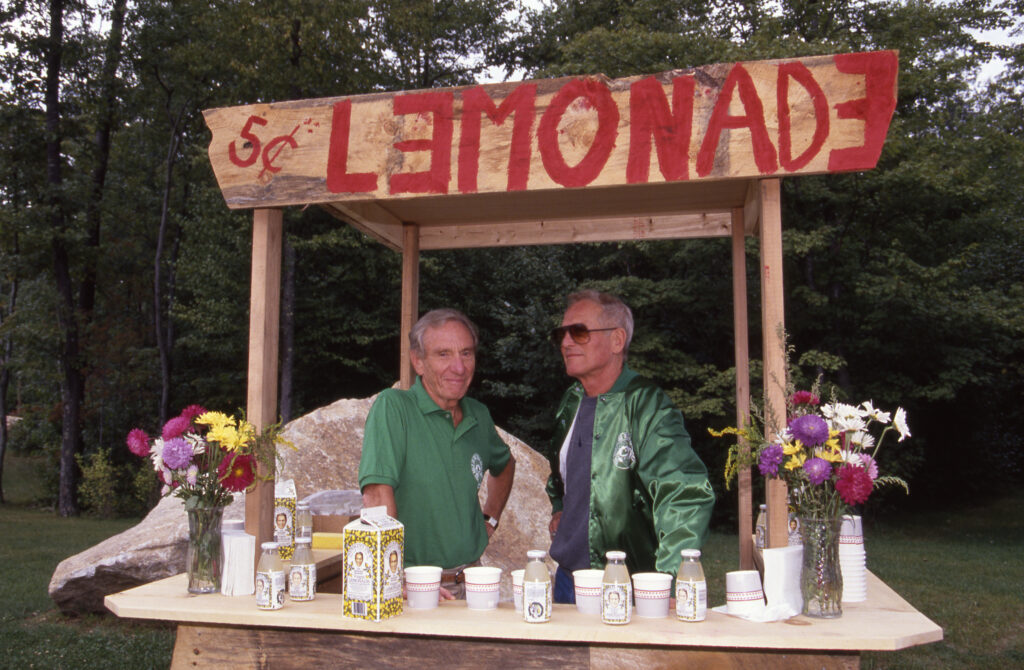 Two men stand at a wooden lemonade stand labeled 5¢ LEMONADE, surrounded by flowers, cups, and lemonade bottles in a park-like setting with trees in the background.