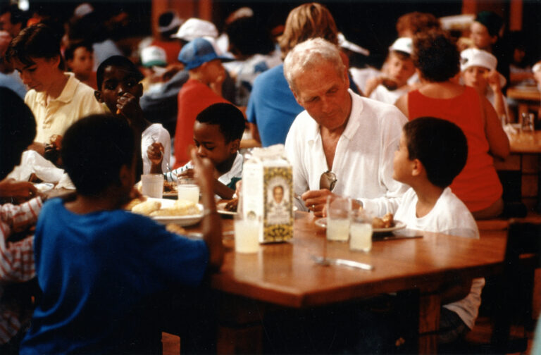 Paul Newman sits at a table with several children, sharing a meal in a busy cafeteria. Cartons of Newman's Own Lemonade and plates of food are on the table, and many people are visible in the background.