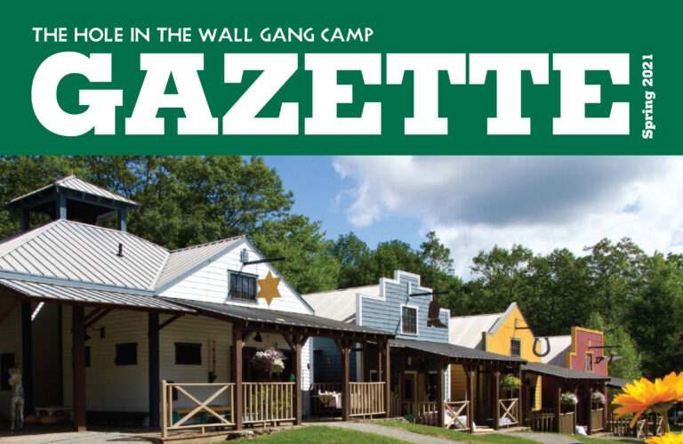 A row of colorful, Western-style buildings under a blue sky, with The Hole in the Wall Gang Camp Gazette Spring 2021 written at the top in bold white letters on a green background.