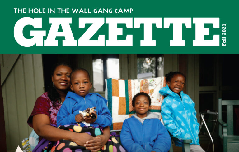 A woman sits on a porch with three smiling children, two wearing blue jackets and one holding a toy horse. A quilt is draped over the chair behind them. The top reads GAZETTE Fall 2021.