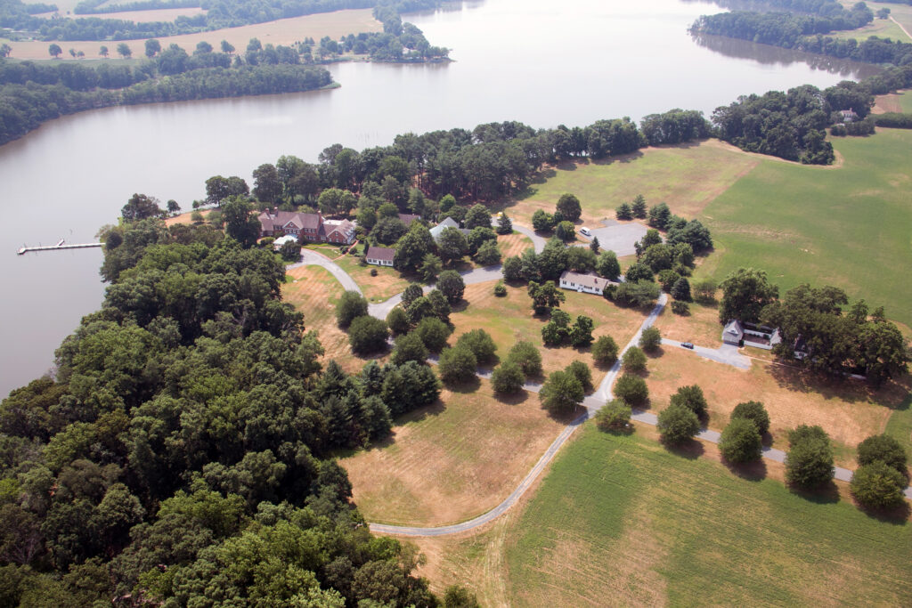 Aerial view of a rural landscape with houses, trees, winding roads, and open fields next to a calm river or lake under a hazy sky.