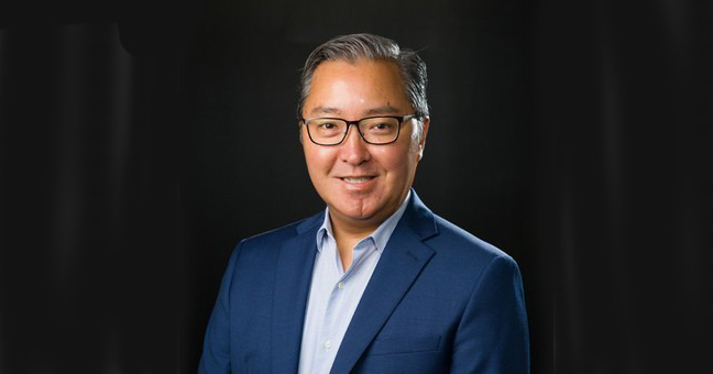 A man with short gray hair, glasses, and a blue suit jacket over a light blue shirt smiles at the camera against a dark background.