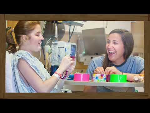 A patient in a hospital gown and an IV sits smiling with another young woman, making colorful crafts together at a table in a hospital room.
