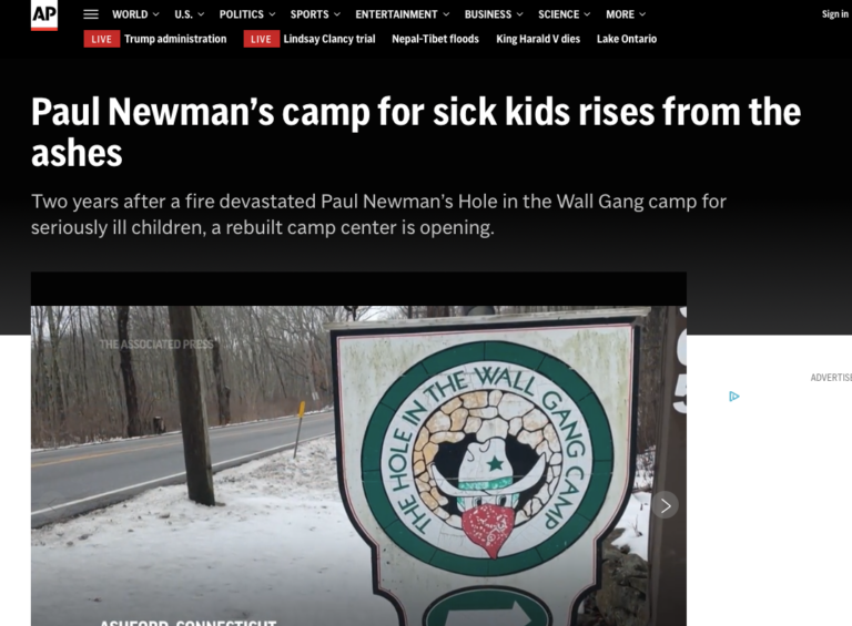 A sign for The Hole in the Wall Gang Camp stands near a road, partially covered in snow, with trees in the background. The sign features a cowboy hat, red bandana, and green sheriffs star.
