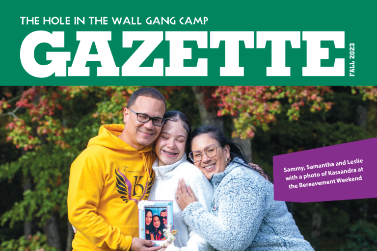 A smiling family of three hugs outdoors, holding a framed photo. Autumn trees are in the background. The image appears on the cover of The Hole in the Wall Gang Camp Gazette Fall 2023 issue.
