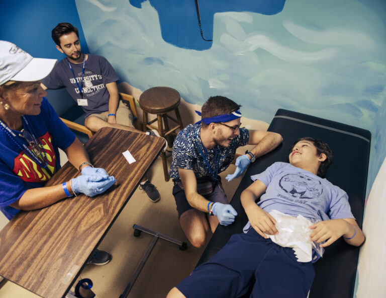 A young person lies on a medical bed with an ice pack on their stomach, while three adults smile and engage with them in a brightly lit room, showing a supportive and friendly atmosphere.