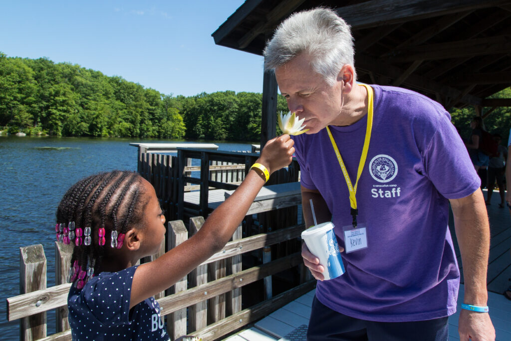 A young girl playfully offers a flower to an older man wearing a purple staff shirt by a lakeside dock on a sunny day.