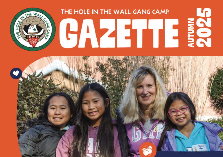 Four people, three children and one adult, smile together outdoors in front of tall grass. The image is the cover of The Hole in the Wall Gang Camp Gazette, Autumn 2025..
