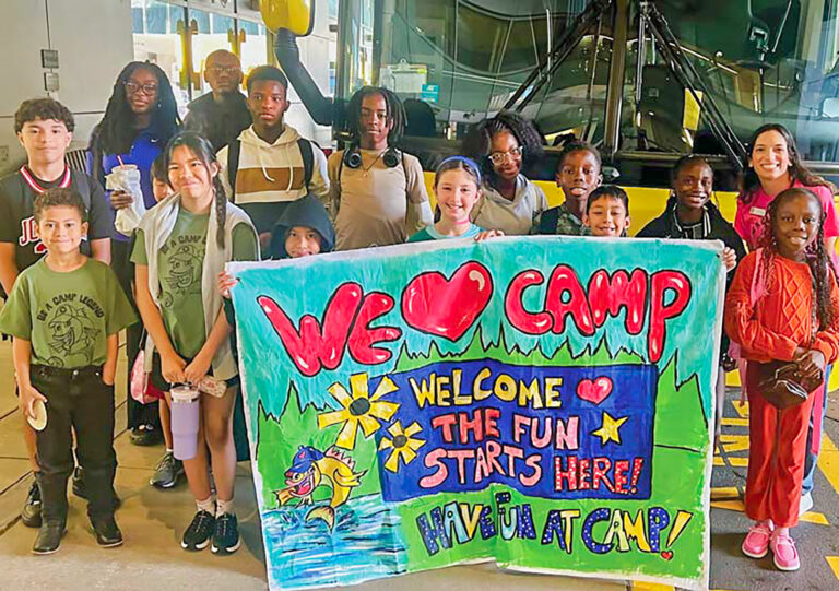 A group of smiling children and adults hold a colorful sign that reads, We ♥ Camp. Welcome. The fun starts here! Have fun at camp! They stand in front of a bus, excited to start their camp adventure.