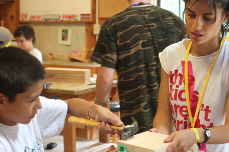 A woman helps a boy use a hammer on a piece of wood in a workshop. Other people and woodworking tools are visible in the background.
