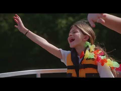 Young girl wearing a yellow life jacket and colorful lei, smiling and reaching out with her arm while enjoying a boat ride outdoors.