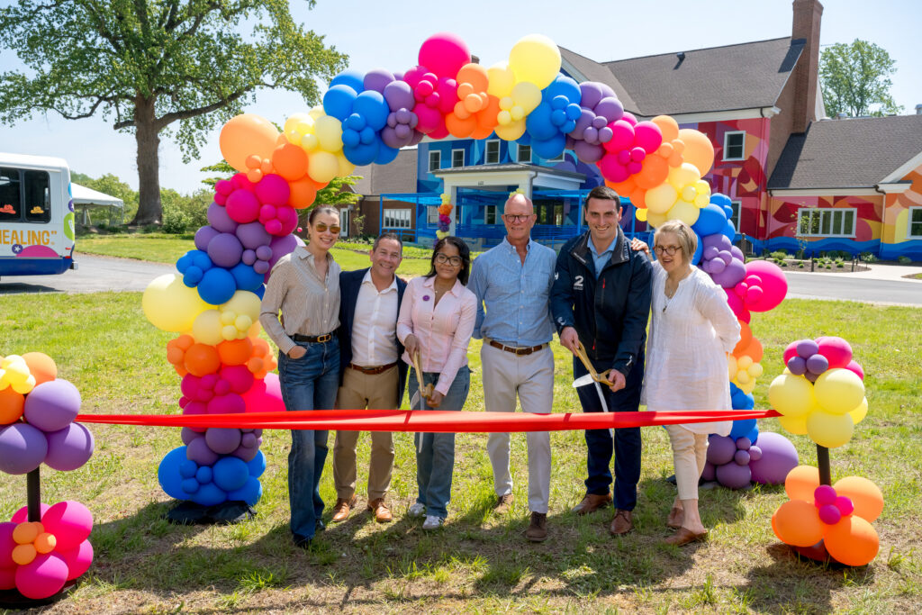 Six people stand smiling under a colorful balloon arch at a ribbon-cutting ceremony outdoors, with scissors and a red ribbon in front; a decorated building and bus are visible in the background.