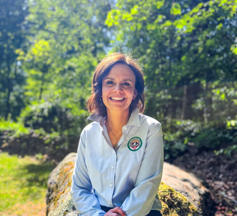 A woman with brown hair, wearing a light blue shirt with a circular embroidered patch, sits on a rock outdoors, smiling, with sunlight and green trees in the background.