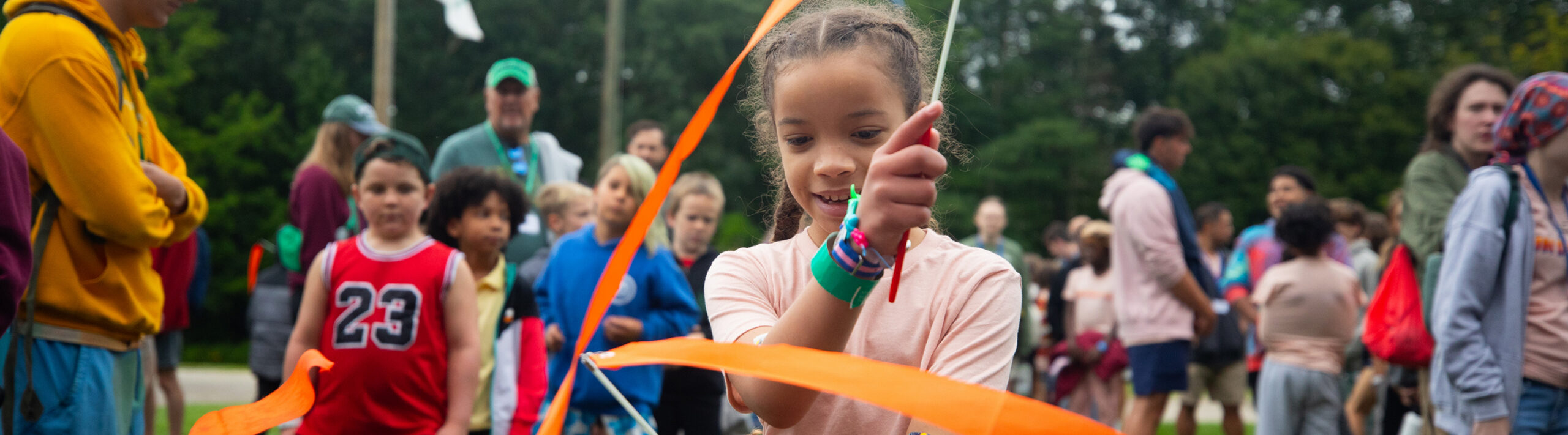A young girl plays with orange streamers outdoors, surrounded by a group of children and adults on a cloudy day.