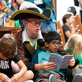 An adult dressed as a pirate reads a book to a group of children, one of whom is sitting on his lap, while other kids sit nearby and listen attentively in a lively indoor setting.