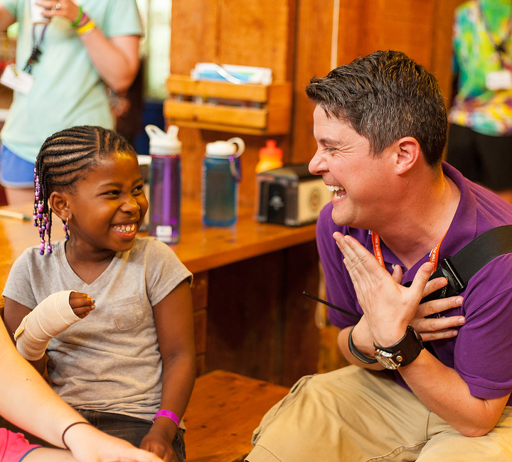 A young girl with braids and a bandaged hand smiles at an adult in a purple shirt. Both are laughing together while sitting at a wooden table in a cheerful indoor setting.