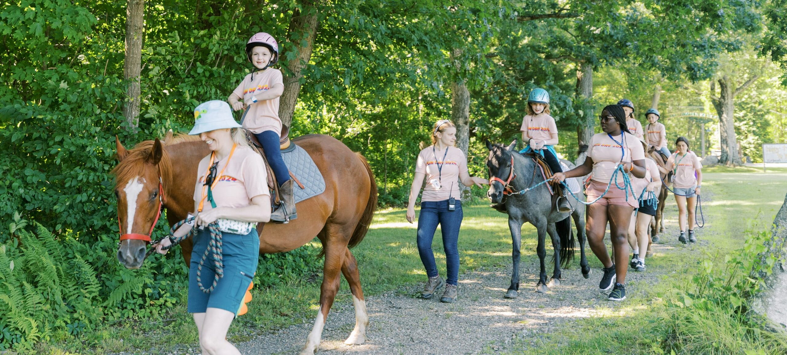 Children wearing helmets ride horses along a forest path, led and guided by adults walking beside them. Everyone wears matching light shirts, and the scene is bright and cheerful. Lush greenery surrounds the group.