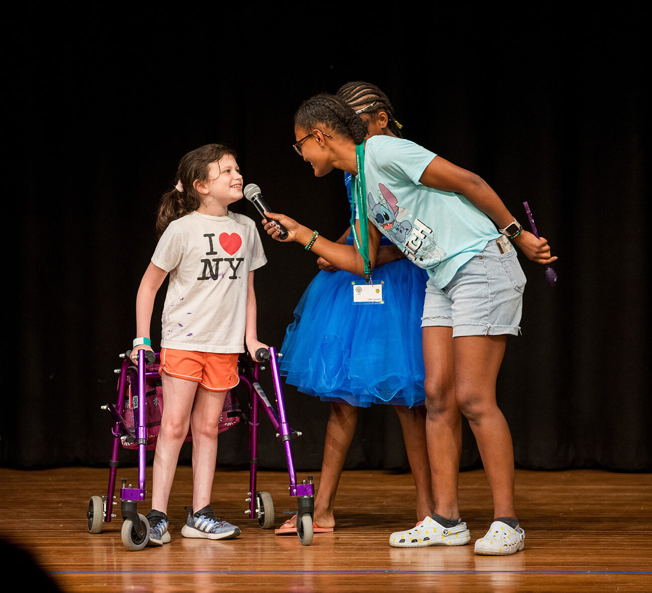 A young girl with a walker smiles as another girl holds a microphone for her on stage. A third girl in a blue dress stands behind them. All appear happy, performing or participating in an event.
