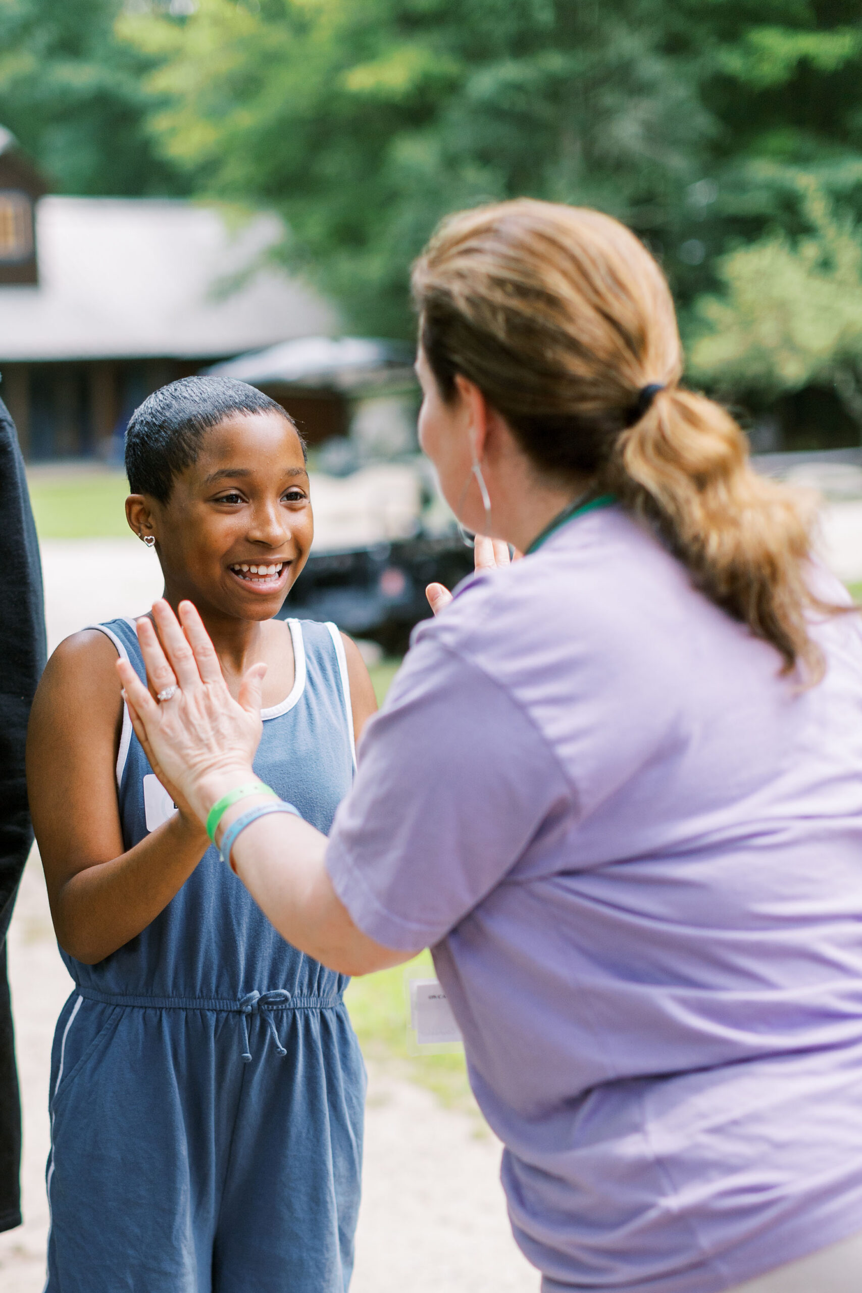 A smiling child in a blue romper gives a high five to an adult in a lavender shirt outdoors, with green trees and a building in the blurred background.