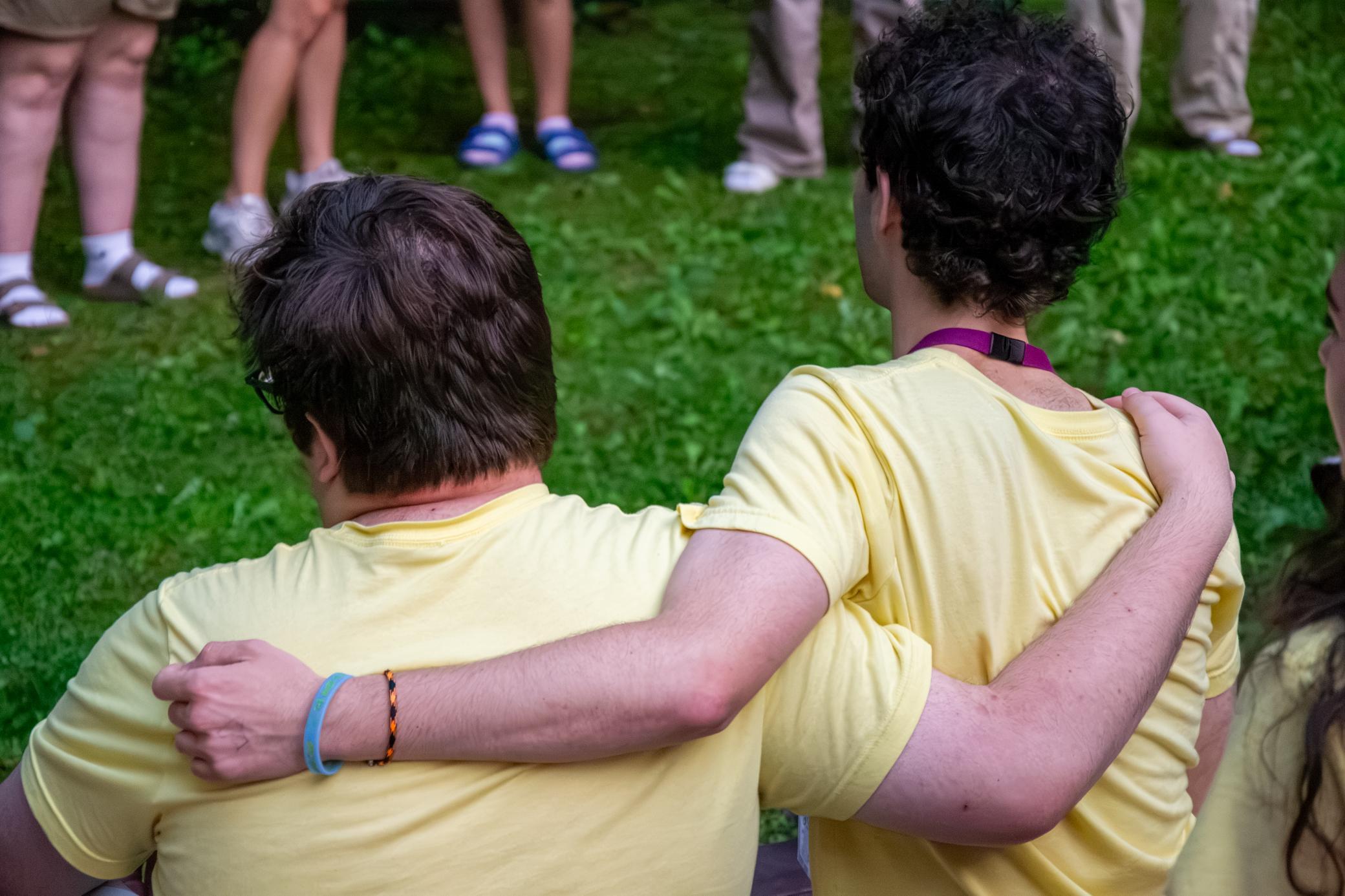 Two people wearing yellow shirts sit on grass, one with an arm around the others shoulders. Other people, some standing and some sitting, are visible in the background.