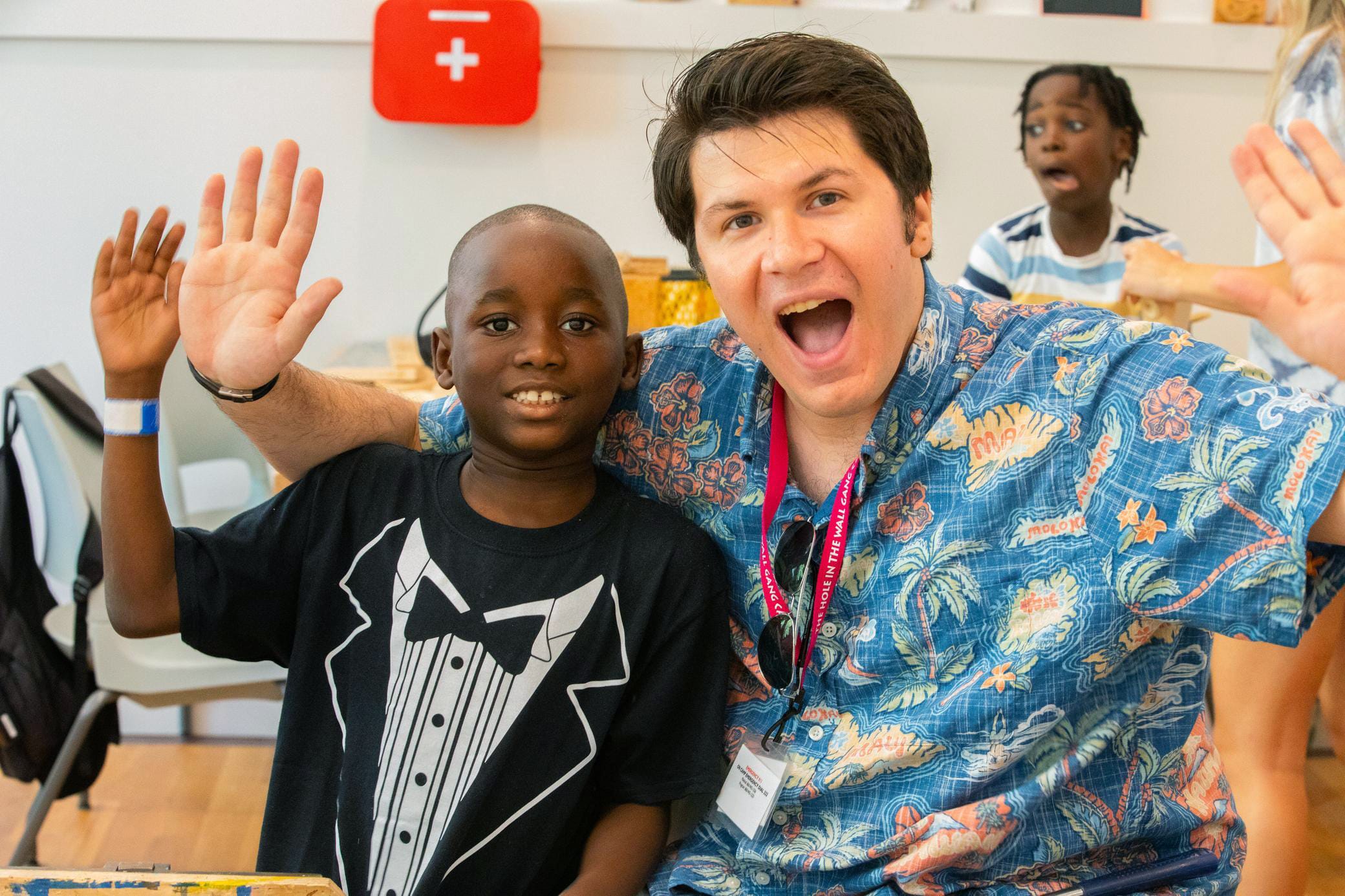 A smiling adult and child sit together, waving at the camera. The child wears a tuxedo-print shirt. A surprised child is in the background. Both appear happy and engaged in a cheerful, indoor setting.