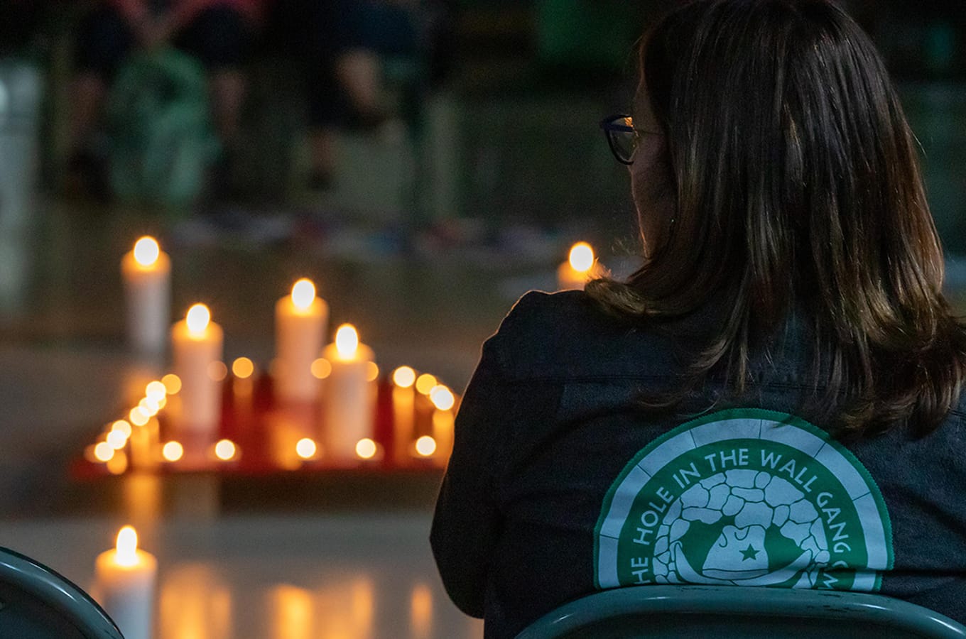 A person sits facing an arrangement of lit candles. They wear a dark jacket with The Hole in the Wall Gang Camp logo on the back. The setting appears quiet and reflective.