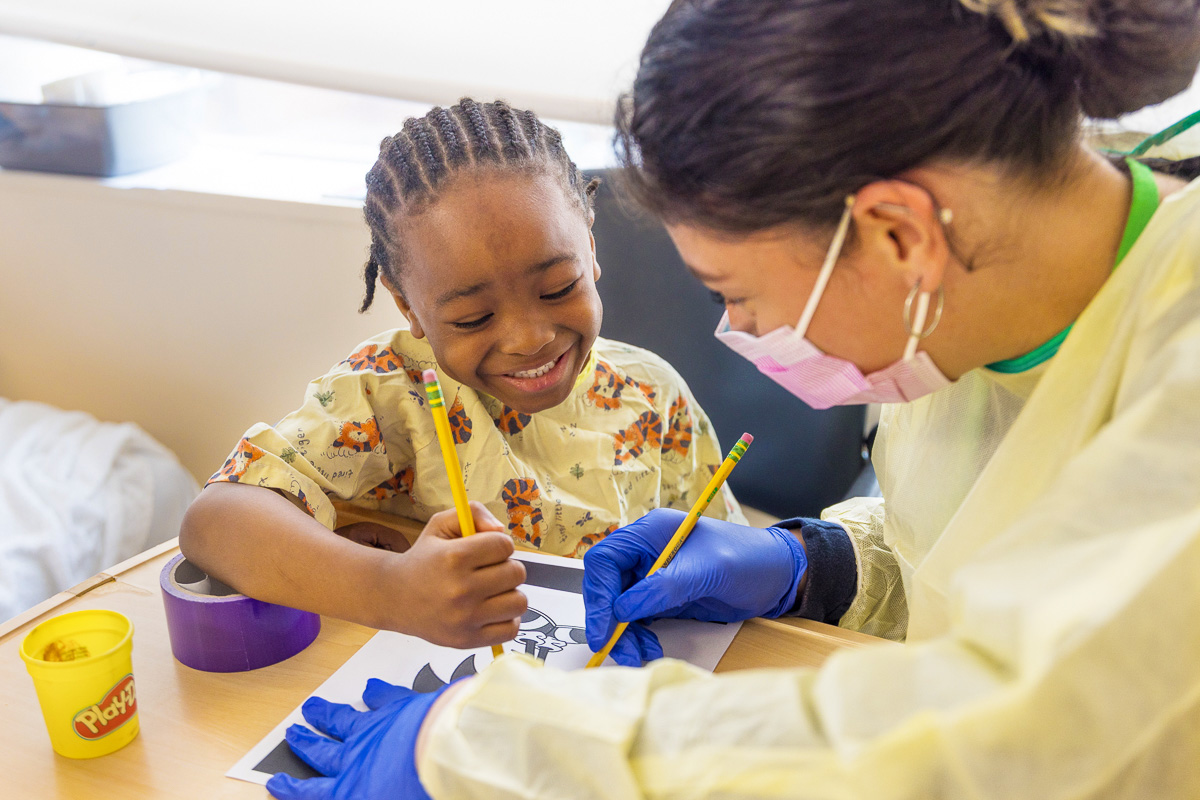 A child and an adult wearing gloves and a mask smile while coloring with pencils at a table. The child wears a hospital gown and there is a Play-Doh container nearby.