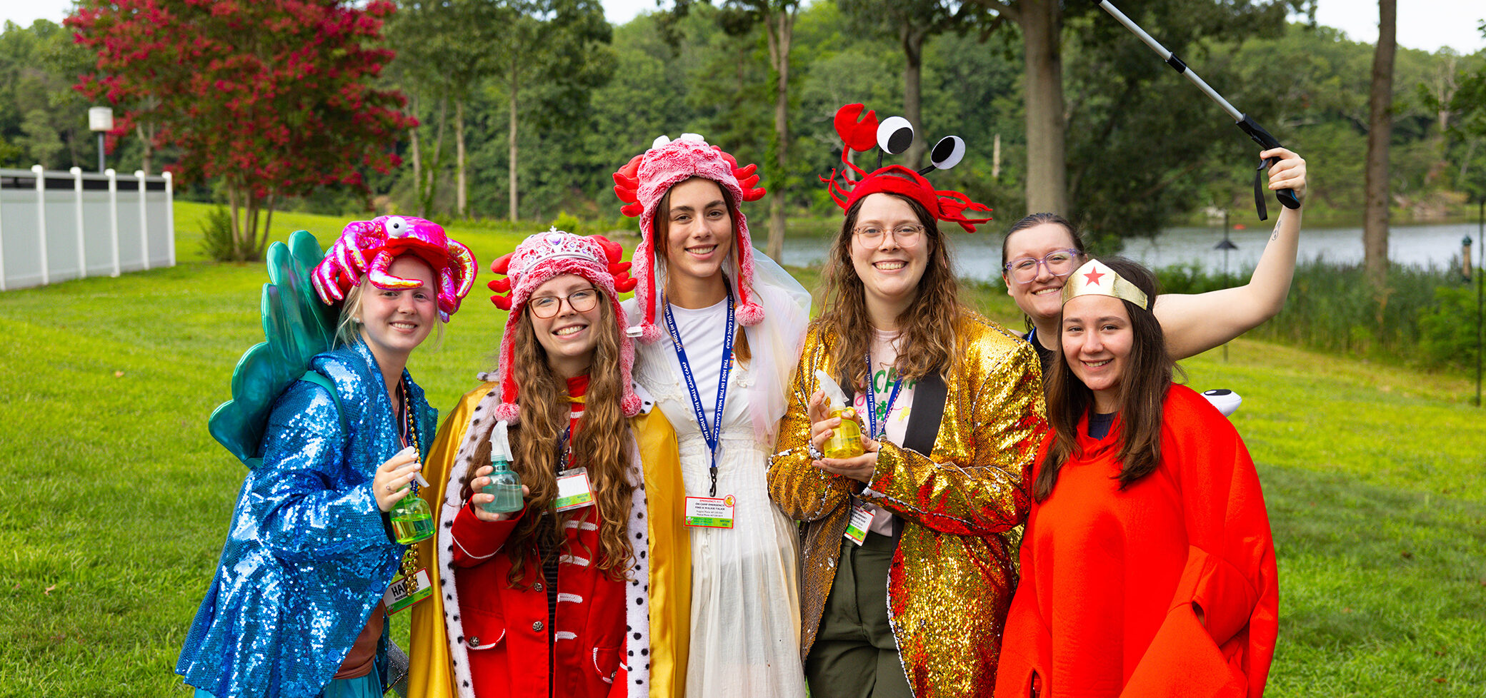 Six people in colorful, whimsical costumes stand together outside on green grass, smiling at the camera. Trees and a pond are in the background, and one person holds a selfie stick.