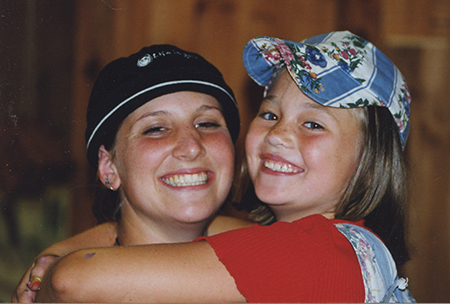 Two people smiling and hugging. The adult on the left wears a black hat, while the child on the right wears a colorful cap, a red shirt, and overalls. Both appear happy in a warmly lit indoor setting.