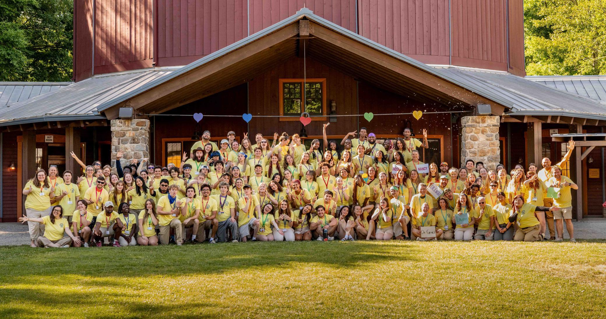 A large group of people in yellow shirts pose together in front of a rustic building with a lawn and trees in the background, smiling and celebrating.