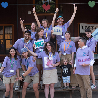 A group of people in matching purple shirts pose playfully on picnic tables, some holding signs with names. Colorful heart shapes hang above them. A child stands in front, and a building is visible in the background.